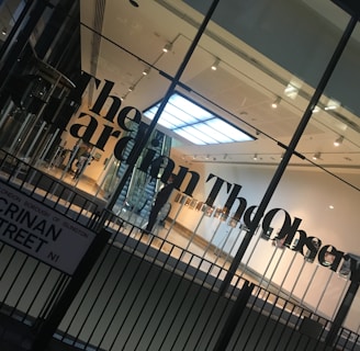 A modern glass building entrance with large black lettering spelling out a newspaper name on the glass. The interior appears well-lit with a visible staircase, minimalistic design, and a sign with a street name in the foreground.