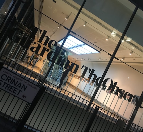 A modern glass building entrance with large black lettering spelling out a newspaper name on the glass. The interior appears well-lit with a visible staircase, minimalistic design, and a sign with a street name in the foreground.